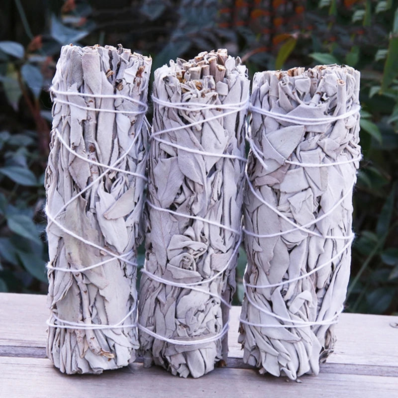 Three white sage bundles standing upright on a wooden surface, wrapped in white string. The natural texture of the dried leaves is visible, with a garden in the background suggesting a natural setting.