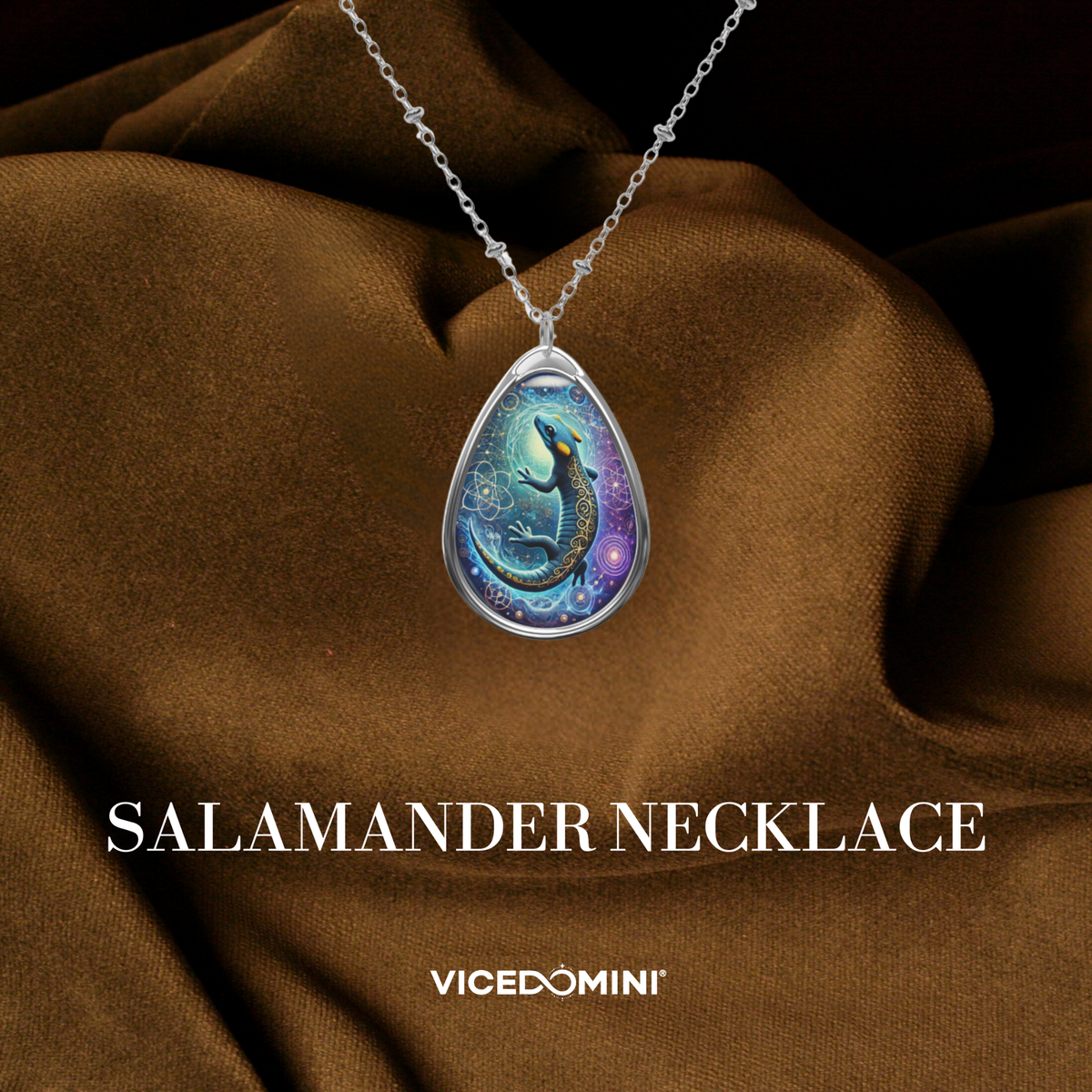 A teardrop-shaped pendant necklace with an intricate illustration of a glowing blue and black salamander surrounded by celestial and geometric patterns. The necklace is displayed on rich brown fabric with "SALAMANDER NECKLACE" and "VICEDOMINI" in white text