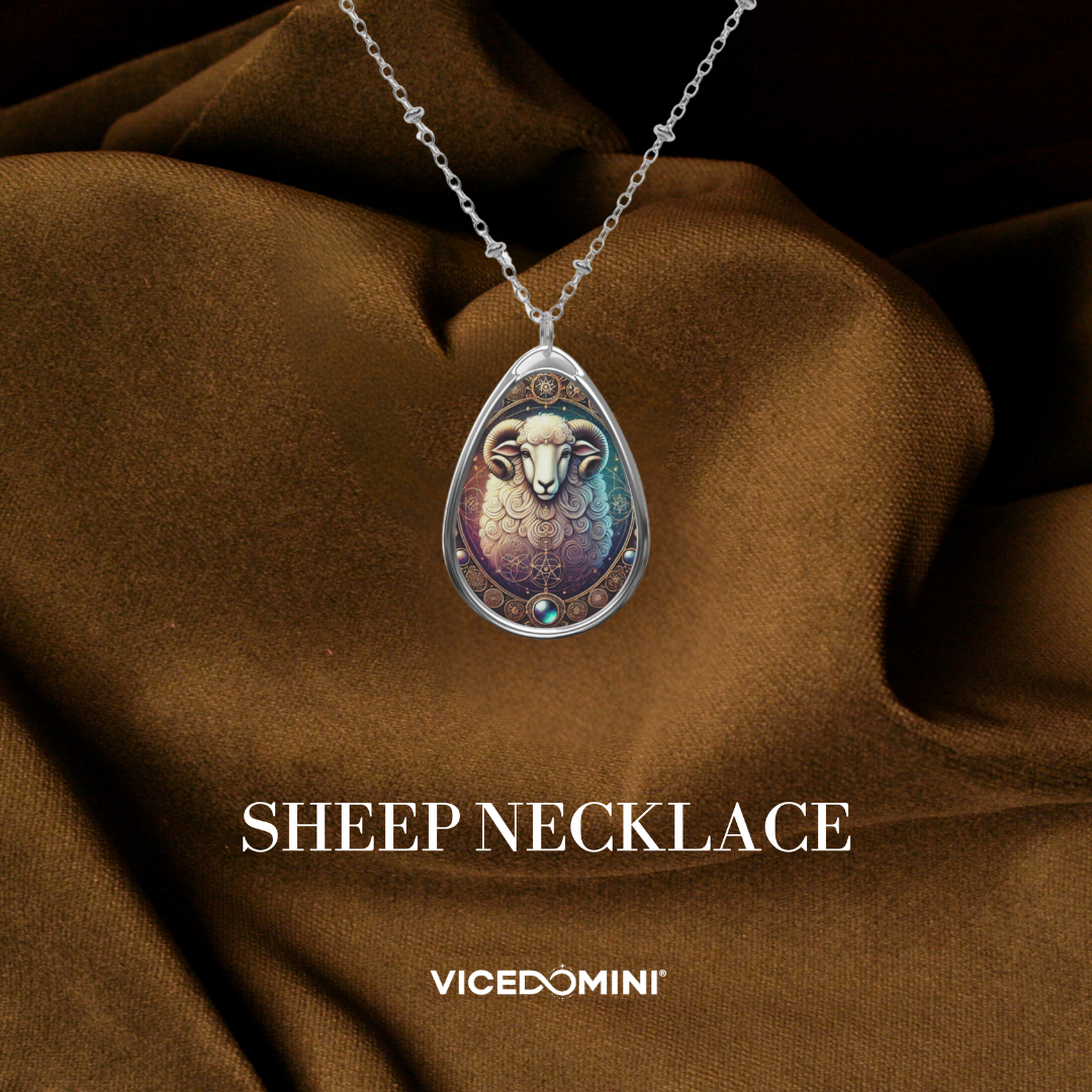 A close-up of a teardrop-shaped pendant necklace featuring an intricately designed sheep head with curled horns. The pendant has ornate golden details and a gemstone accent, set against a rich brown fabric backdrop.