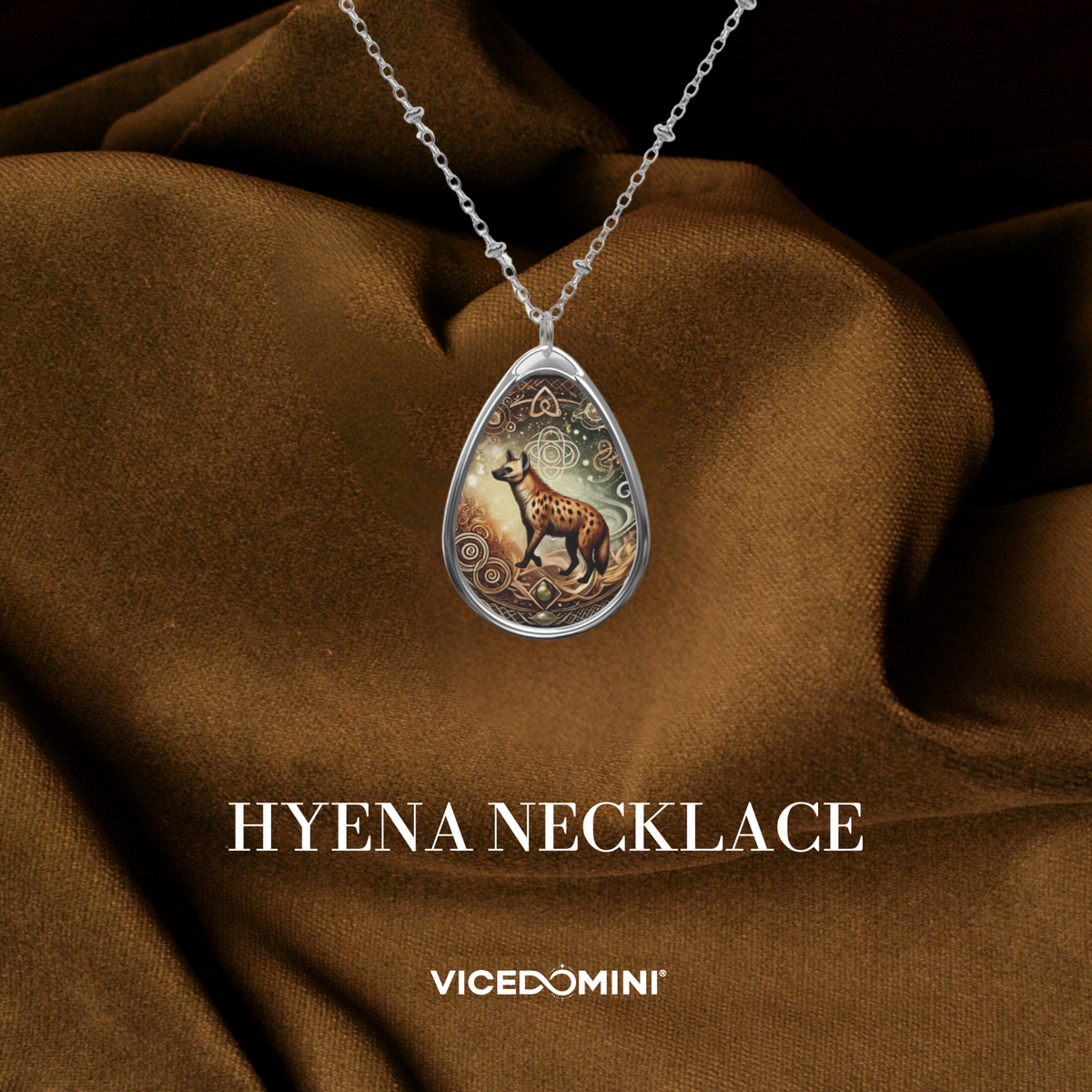 A teardrop-shaped silver pendant necklace featuring an illustrated hyena on a patterned background, draped over brown satin fabric. The text 'HYENA NECKLACE' and 'VICEDOMINI' are displayed below.