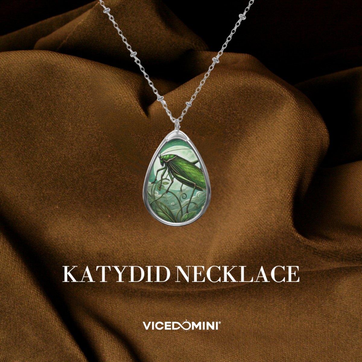 A silver teardrop pendant necklace featuring a green katydid insect illustration, draped over brown satin fabric with the text 'KATYDID NECKLACE' and 'VICEDOMINI' below.