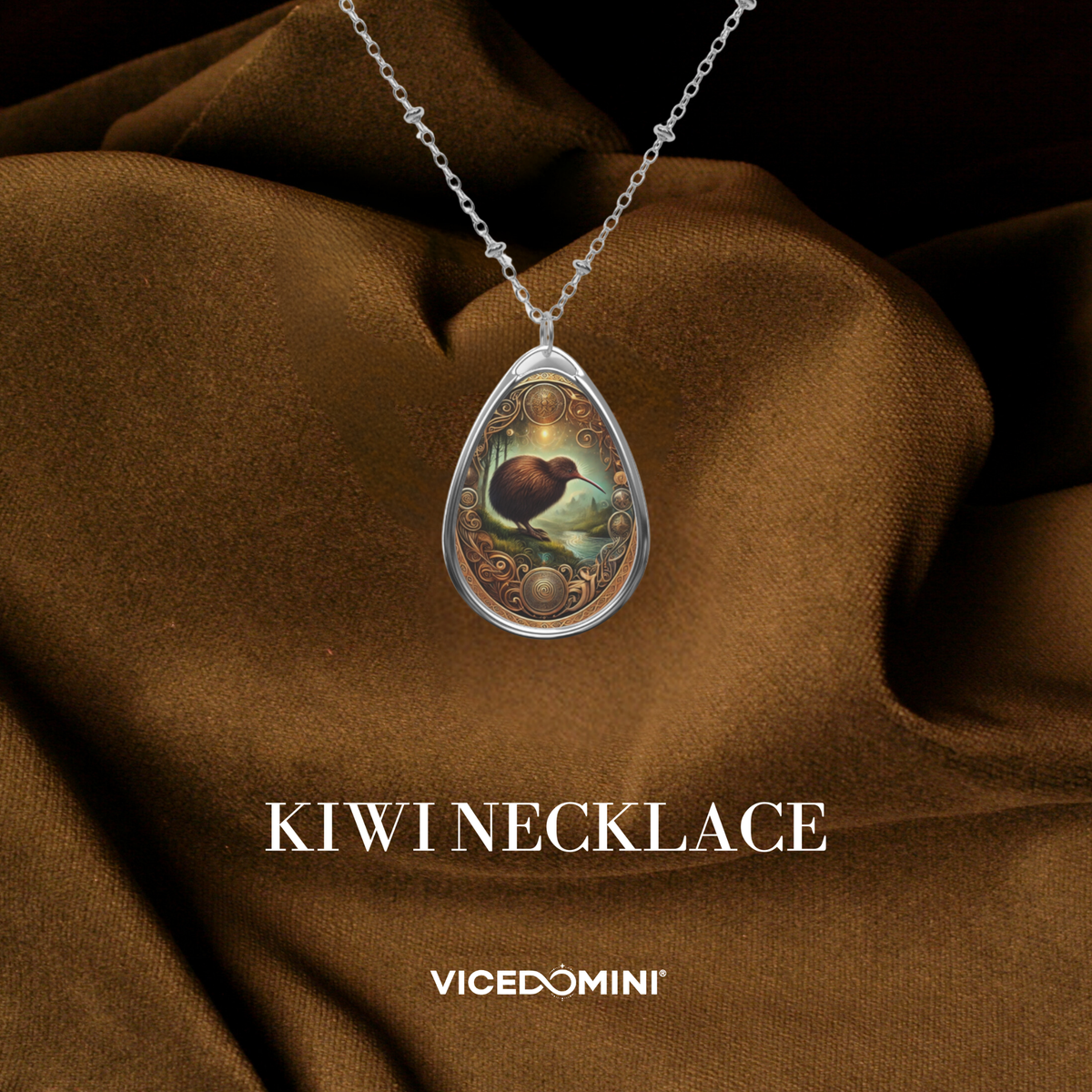 A teardrop-shaped pendant necklace featuring a detailed kiwi bird illustration in a silver frame, draped over brown satin fabric with the text 'KIWI NECKLACE' and 'VICEDOMINI' below.