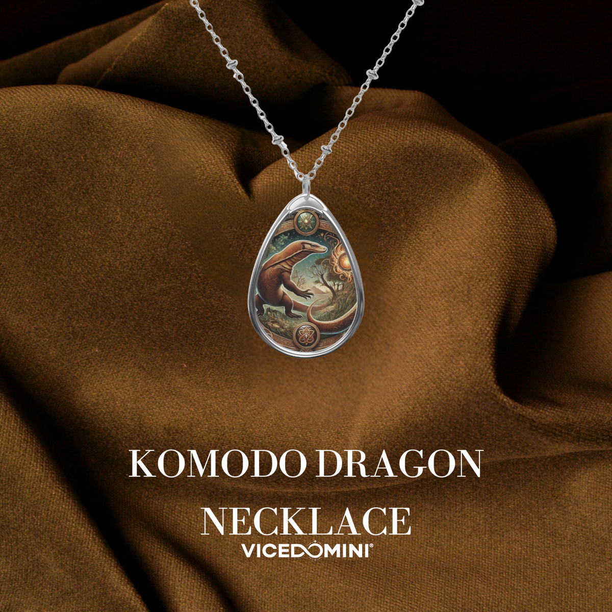 A teardrop-shaped silver pendant necklace featuring an illustrated Komodo dragon design, displayed on draped brown fabric with text reading 'KOMODO DRAGON NECKLACE' and 'VICEDOMINI'.