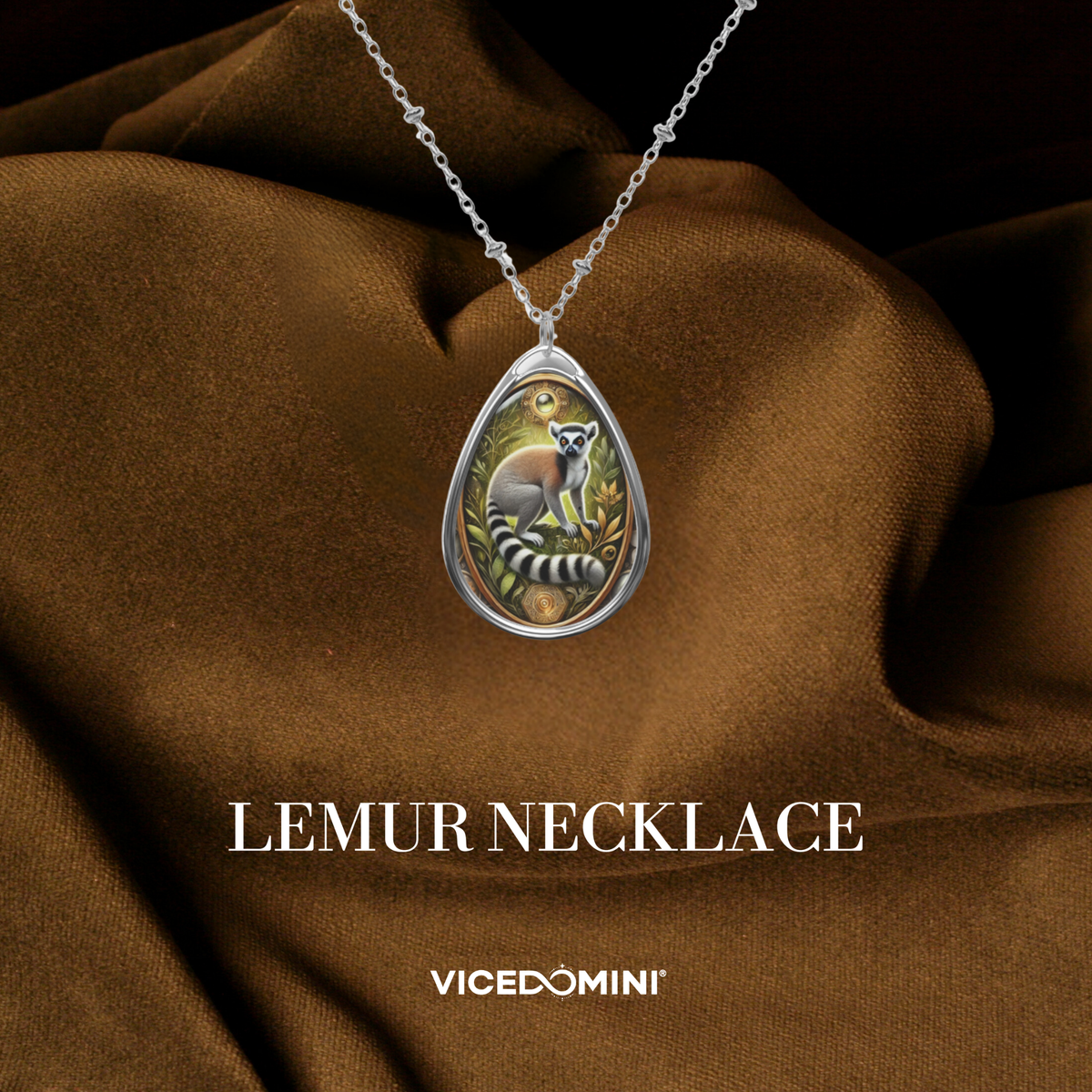 A teardrop-shaped silver pendant necklace featuring a colorful illustration of a lemur, displayed on draped brown fabric with the text 'LEMUR NECKLACE' and 'VICEDOMINI' below.