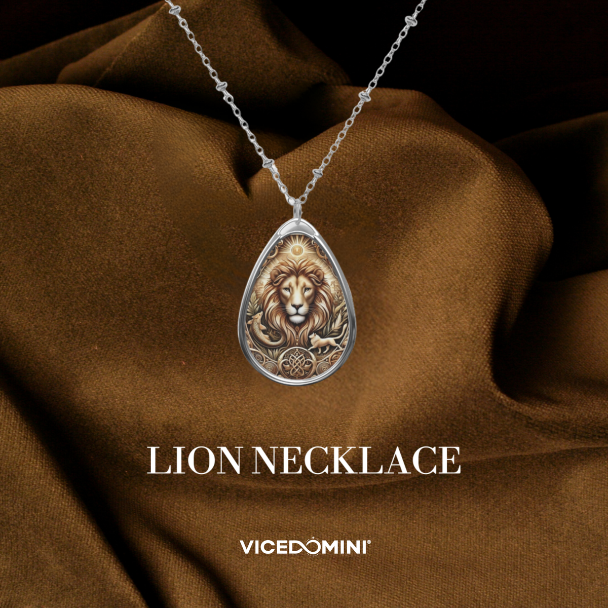 A silver teardrop pendant necklace featuring an embossed lion design, draped over brown satin fabric with the text 'LION NECKLACE' and 'VICEDOMINI' below.