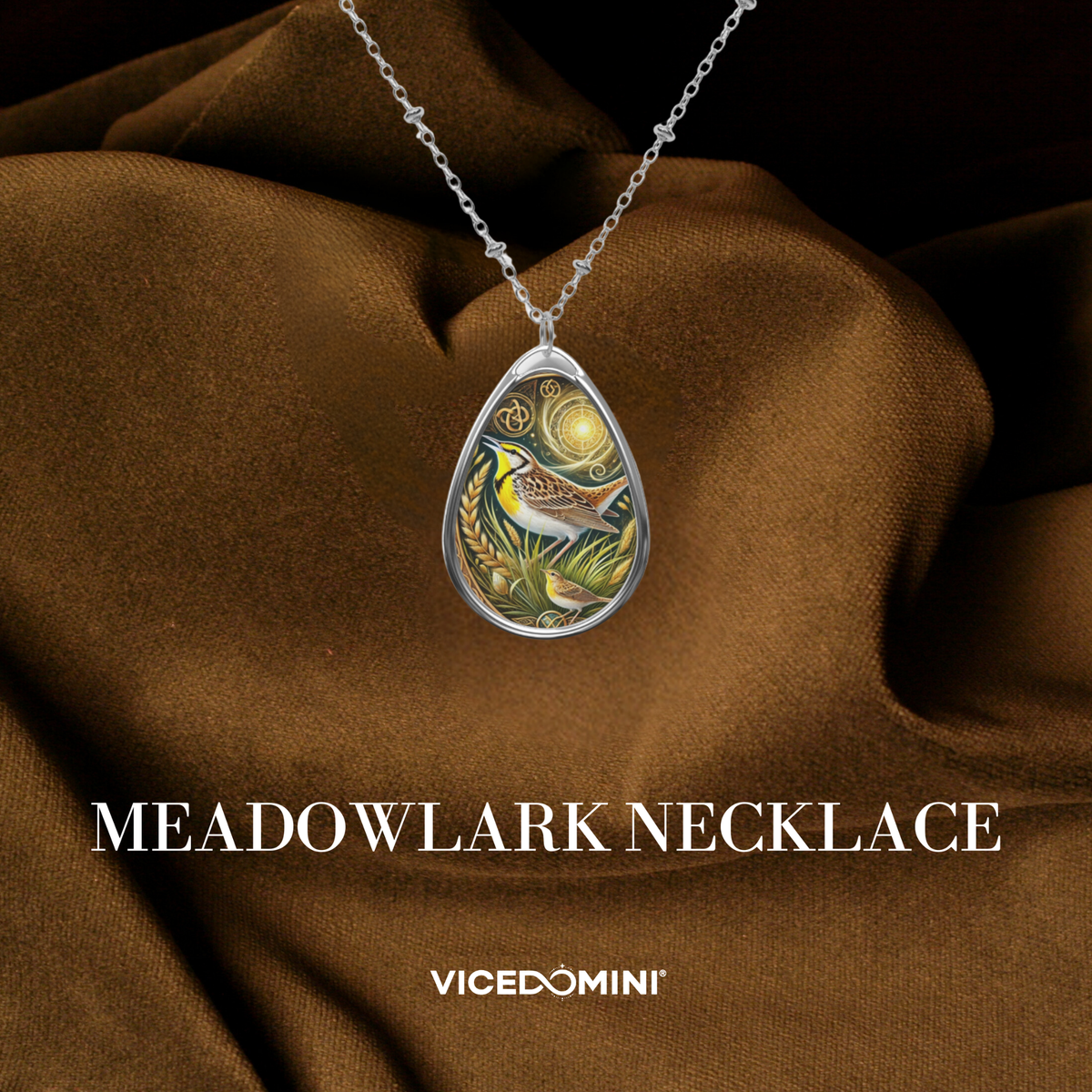 A teardrop-shaped silver pendant necklace featuring a colorful illustration of a bird in a marsh under a full moon, draped over brown satin fabric. The text 'MEADOWLARK NECKLACE' and the brand 'VICEDOMINI' are displayed below.