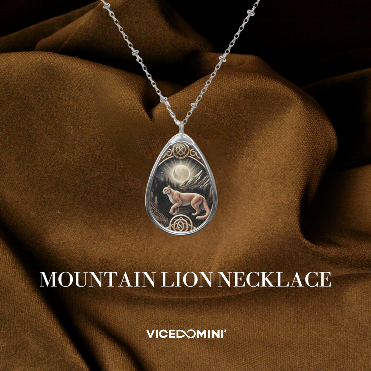 A silver teardrop pendant necklace featuring an illustrated mountain lion under a full moon, draped over brown satin fabric. The text 'MOUNTAIN LION NECKLACE' and the brand 'VICEDOMINI' are displayed below.
