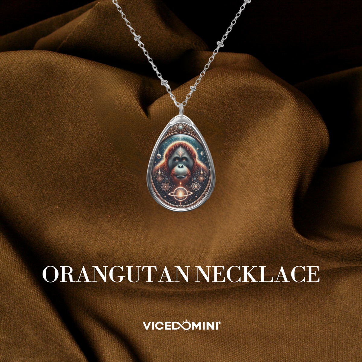 A teardrop-shaped silver pendant necklace featuring a colorful illustration of an orangutan, displayed on draped brown fabric with the text 'ORANGUTAN NECKLACE' and the brand 'VICEDOMINI' below.