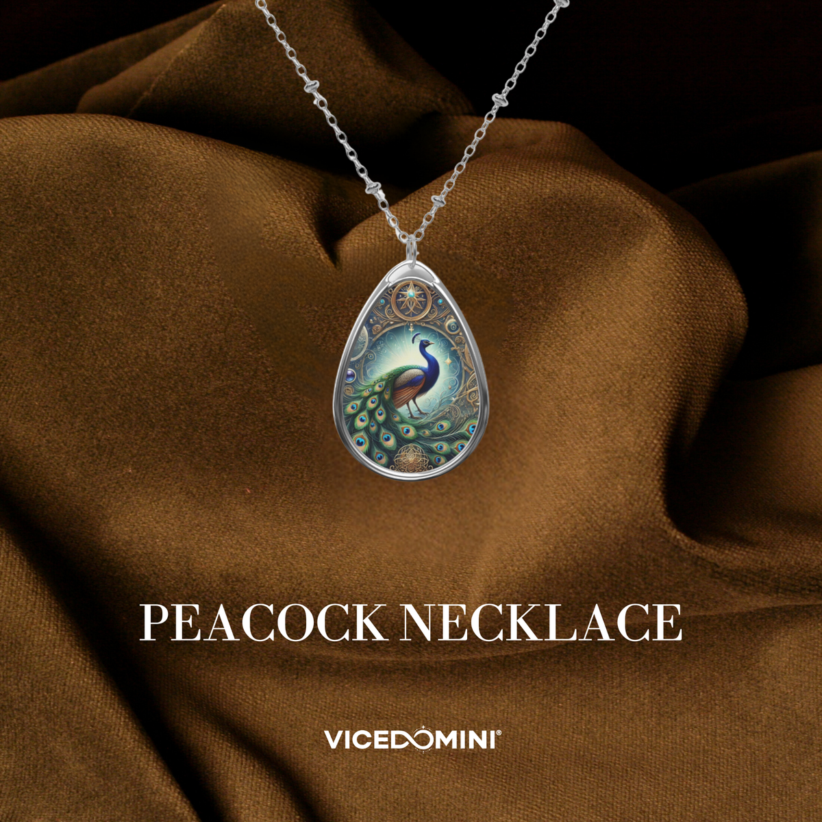 A teardrop-shaped silver pendant necklace featuring a colorful peacock illustration, displayed on draped brown satin fabric with the text 'PEACOCK NECKLACE' and 'VICEDOMINI' below.