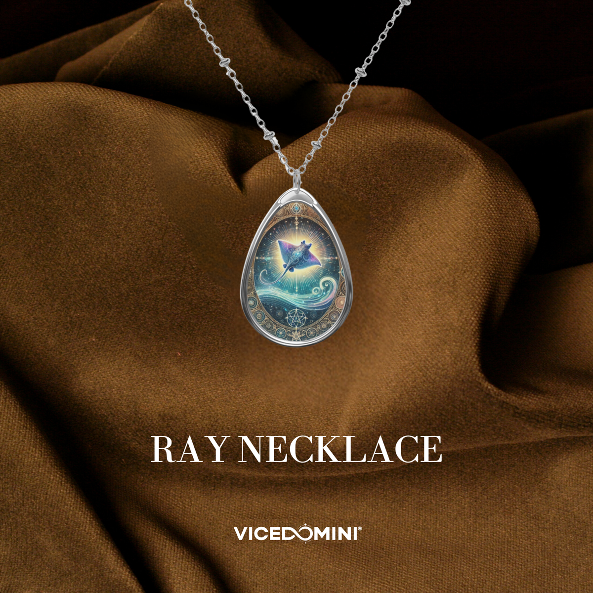 A teardrop-shaped pendant featuring a glowing, celestial-inspired stingray design, surrounded by intricate golden filigree and swirling ocean waves. The pendant hangs from a delicate silver chain, displayed on a luxurious brown fabric background. The text "RAY NECKLACE" and "vicedomini" are printed below.