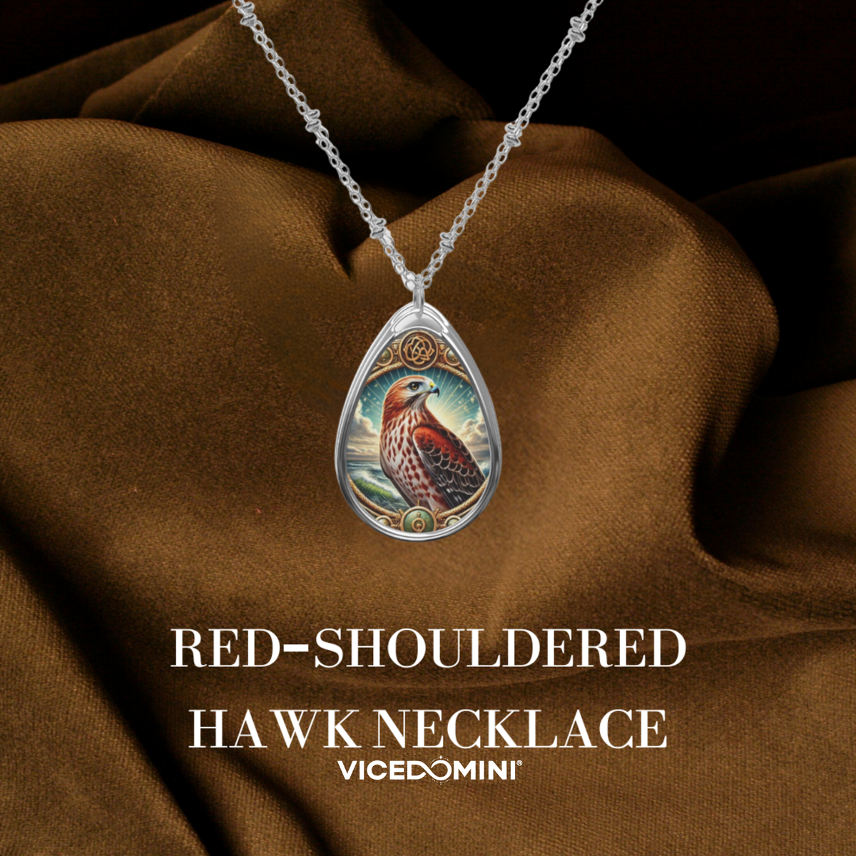 A teardrop-shaped pendant necklace featuring an illustrated red-shouldered hawk on a silver-toned chain, draped over brown satin fabric with the text 'RED-SHOULDERED HAWK NECKLACE' and 'VICEDOMINI' below.