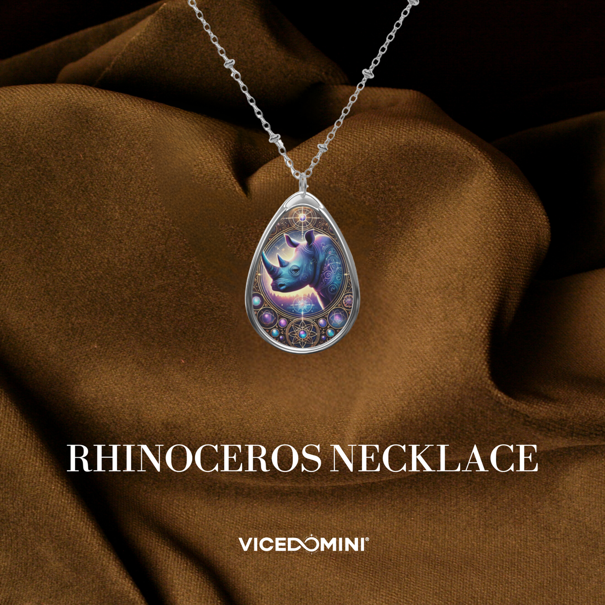 A silver teardrop-shaped pendant necklace featuring a beautifully detailed rhinoceros illustration with mystical purple and blue hues, adorned with intricate geometric designs. The necklace rests on a luxurious brown fabric background with "Rhinoceros Necklace" and "VICEDOMINI" text.