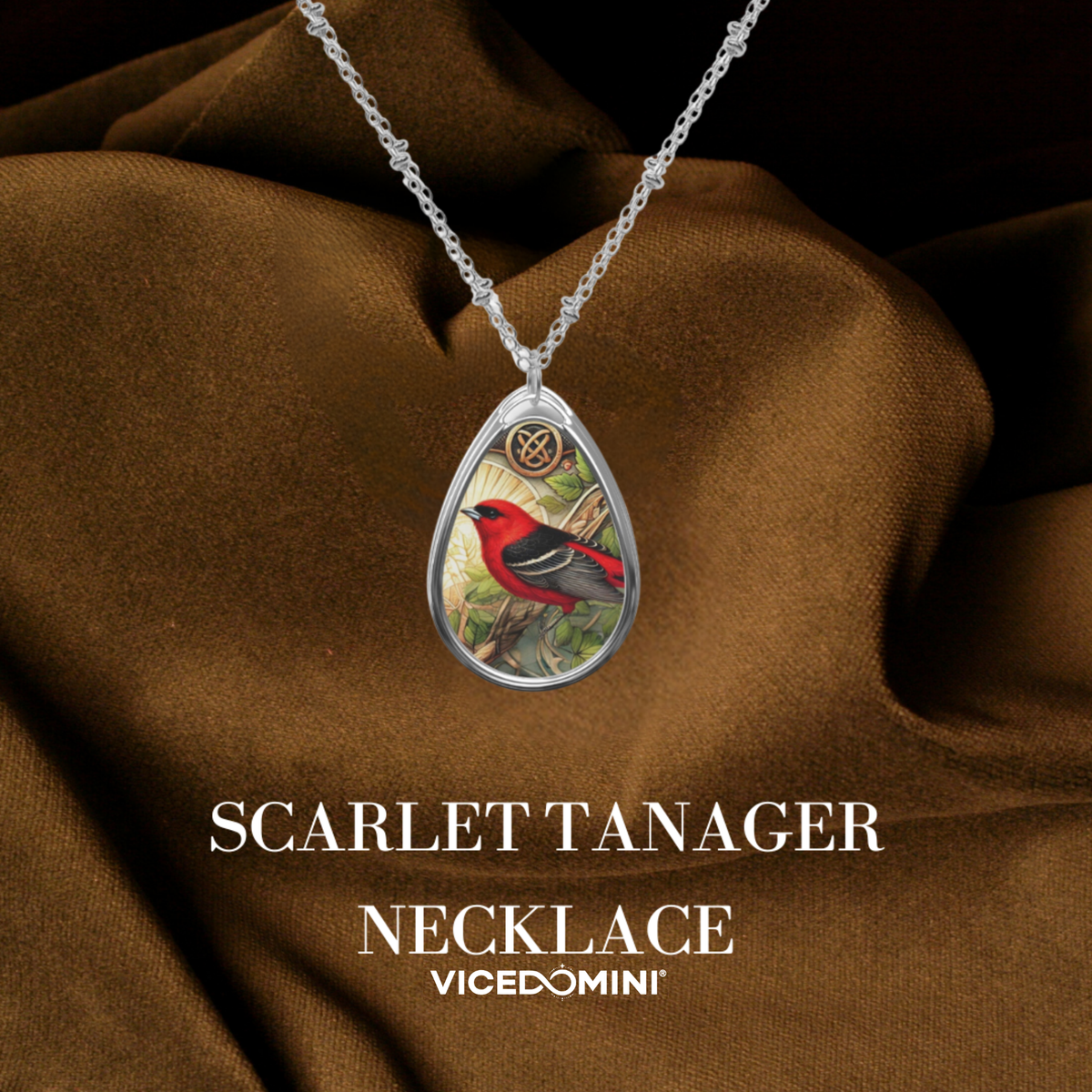 Silver teardrop pendant necklace featuring a detailed illustration of a scarlet tanager bird, displayed on draped brown satin fabric with text overlay 'SCARLET TANAGER NECKLACE' and 'VICEDOMINI'.
