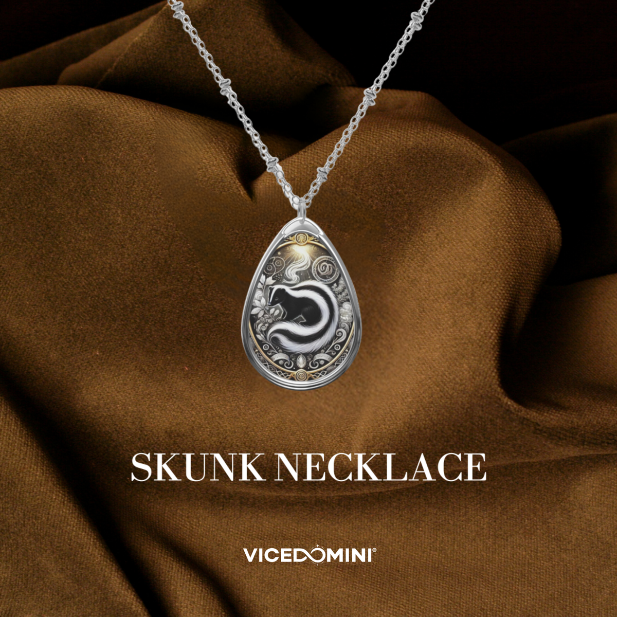 A silver pendant necklace with a teardrop-shaped silver medallion featuring a black and white skunk design, draped over brown satin fabric. The text 'SKUNK NECKLACE' and 'VICEDOMINI' are displayed below.