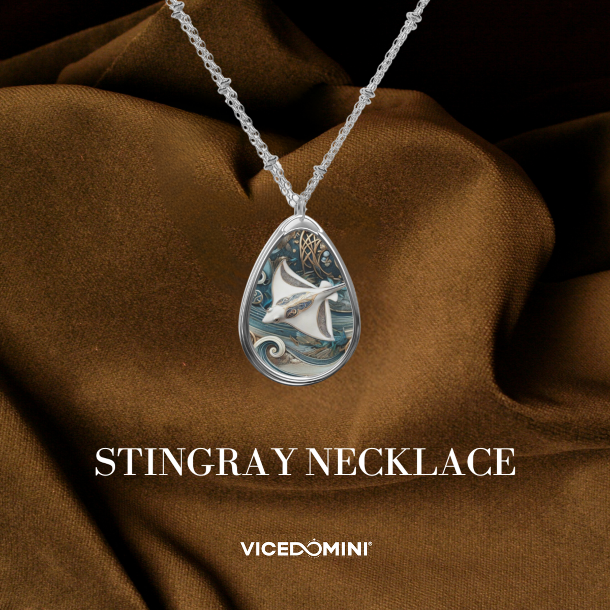 A teardrop-shaped silver pendant necklace featuring a detailed stingray design with blue and white accents, displayed on draped brown fabric with the text 'STINGRAY NECKLACE' and 'VICEDOMINI' below.