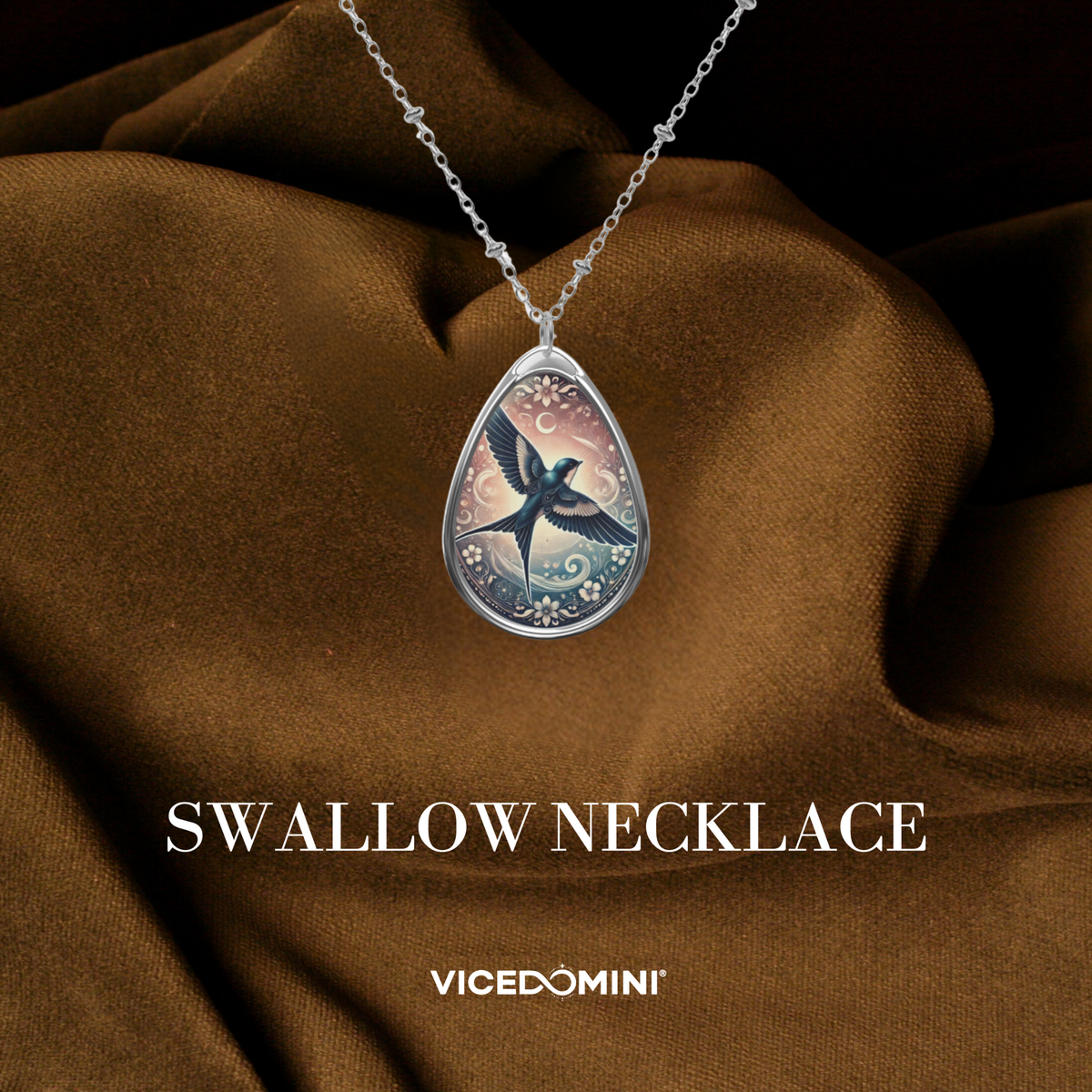 A teardrop-shaped pendant necklace featuring an illustrated penguin in a silver-toned frame, draped over brown satin fabric with the text 'PENGUIN NECKLACE' and 'VICEDOMINI' below.