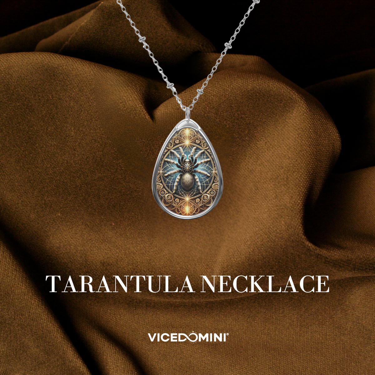 A teardrop-shaped silver pendant necklace featuring an intricate tarantula design with blue, gold, and black details, displayed on draped brown satin fabric. The text 'TARANTULA NECKLACE' and the brand 'VICEDOMINI' are visible at the bottom.
