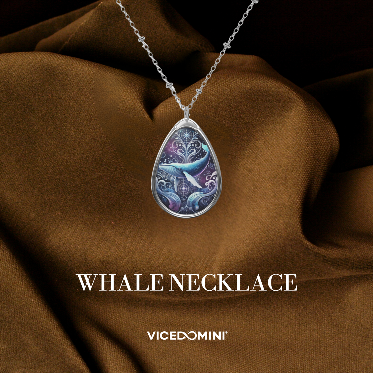 A teardrop-shaped pendant necklace with a detailed whale illustration in shades of blue, purple, and white, set in a silver-toned metal frame and hanging from a silver chain. The pendant is displayed against a draped brown fabric background with the text 'WHALE NECKLACE' and 'VICEDOMINI' below.