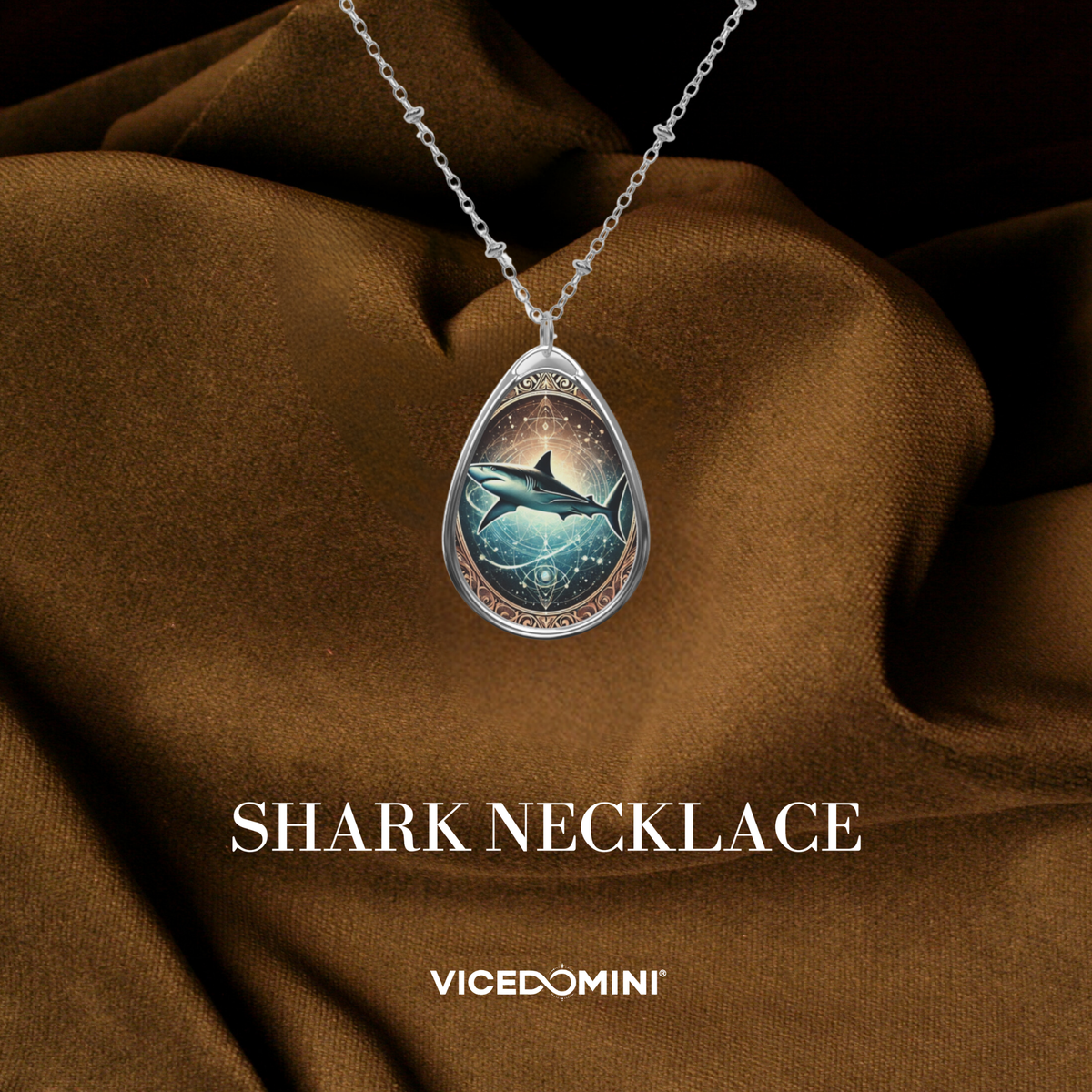A close-up of a teardrop-shaped shark necklace pendant with an intricate celestial design, featuring a shark swimming through a cosmic ocean. The pendant hangs from a delicate silver chain against a textured brown fabric backdrop, with "SHARK NECKLACE" and "VICEDOMINI" text displayed below.