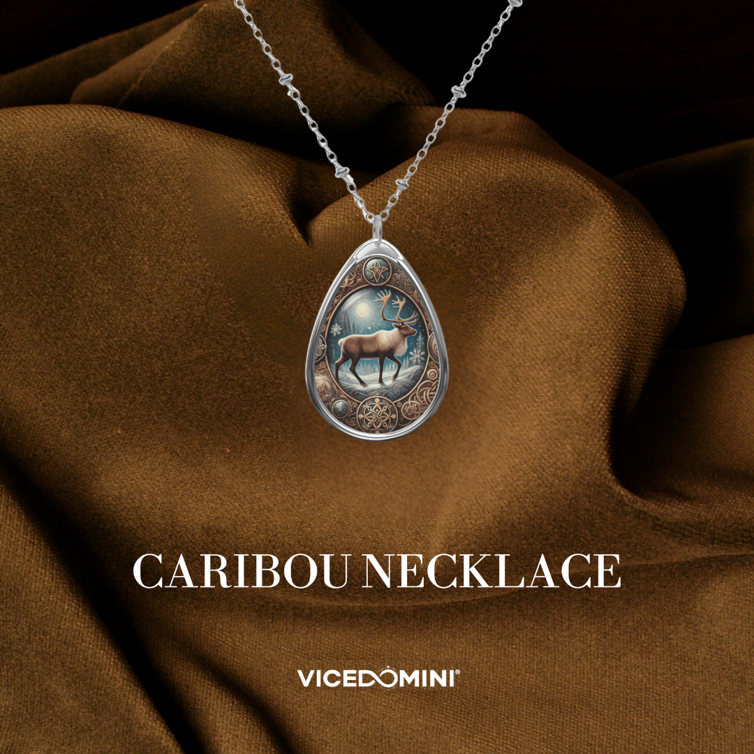 A teardrop-shaped silver pendant necklace featuring an illustrated caribou design, displayed on draped brown fabric with the text 'CARIBOU NECKLACE' and 'VICEDOMINI' below.