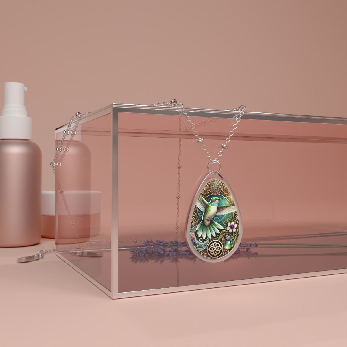 The hummingbird pendant is draped over a transparent glass box, surrounded by soft pink and lavender hues. The intricate details of the pendant’s design, including floral motifs and swirling golden accents, are highlighted in this artistic composition.