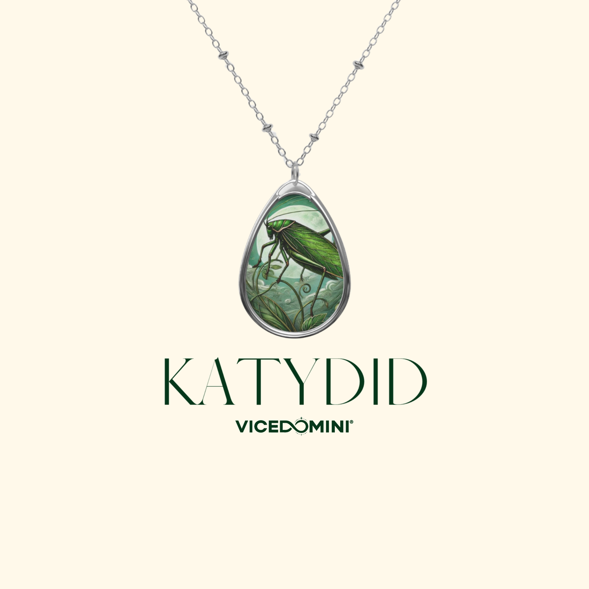 A teardrop-shaped pendant necklace featuring a green dragonfly illustration on a light green background, set in a silver-toned metal frame and suspended from a silver chain. The brand name 'KATYDID' is displayed below the necklace, with 'VICEDOMINI' underneath, all on a solid pale cream background.
