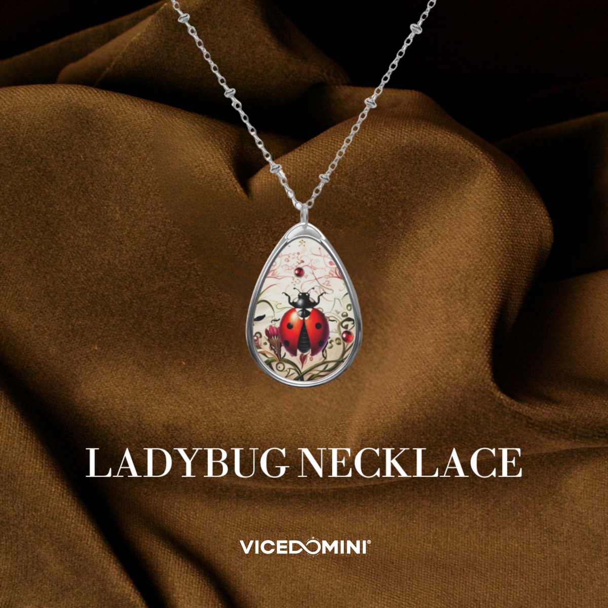 A teardrop-shaped pendant necklace featuring a ladybug illustration on a silver chain, draped over brown satin fabric with the text 'LADYBUG NECKLACE' and 'VICEDOMINI' below.