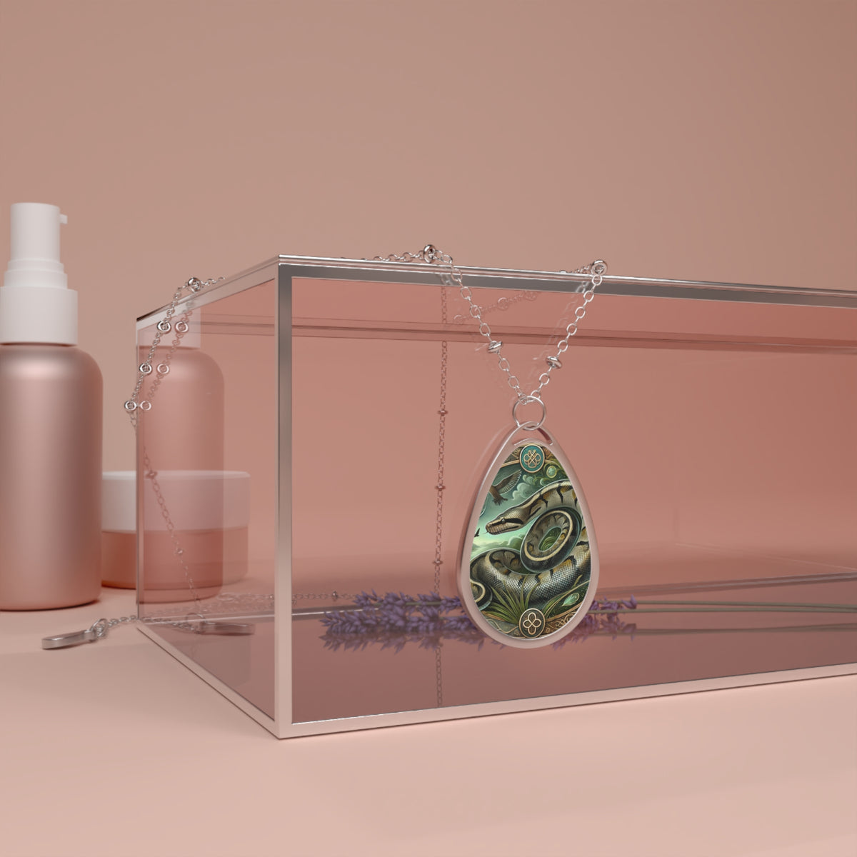 A silver chain necklace with a teardrop-shaped pendant featuring an intricately illustrated anaconda coiled in a lush jungle setting. The pendant is elegantly draped over a transparent jewelry box, surrounded by soft pink beauty product containers and sprigs of lavender in the background.