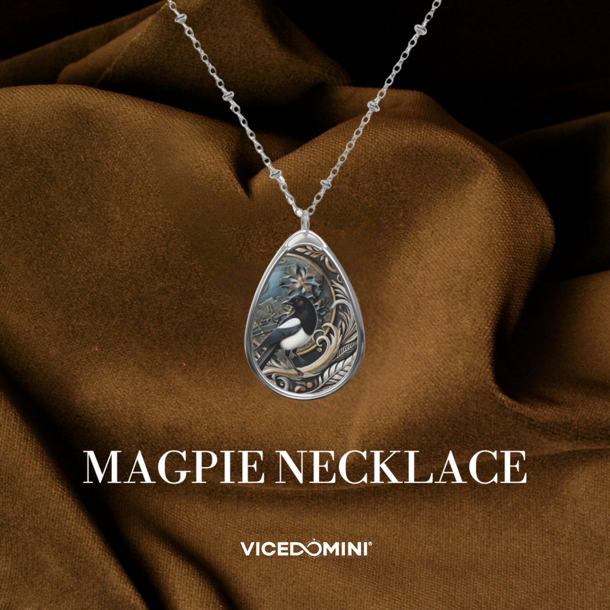 A silver teardrop pendant necklace featuring an engraved magpie bird design, draped over brown satin fabric with the text 'MAGPIE NECKLACE' and 'VICEDOMINI' below.