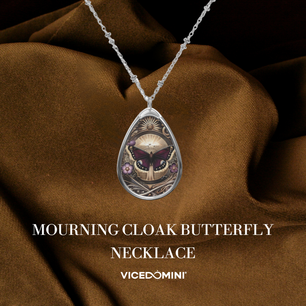 A silver teardrop pendant necklace featuring an intricate illustration of a mourning cloak butterfly, draped over brown satin fabric with text overlay 'MOURNING CLOAK BUTTERFLY NECKLACE' and the brand 'VICEDOMINI'.