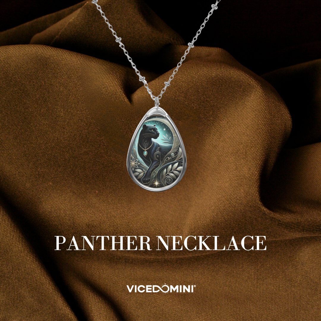 A teardrop-shaped pendant necklace displayed on draped brown satin fabric. The pendant features an intricate silver-toned design with a black panther silhouette, a crescent moon, and a glowing blue-green background.