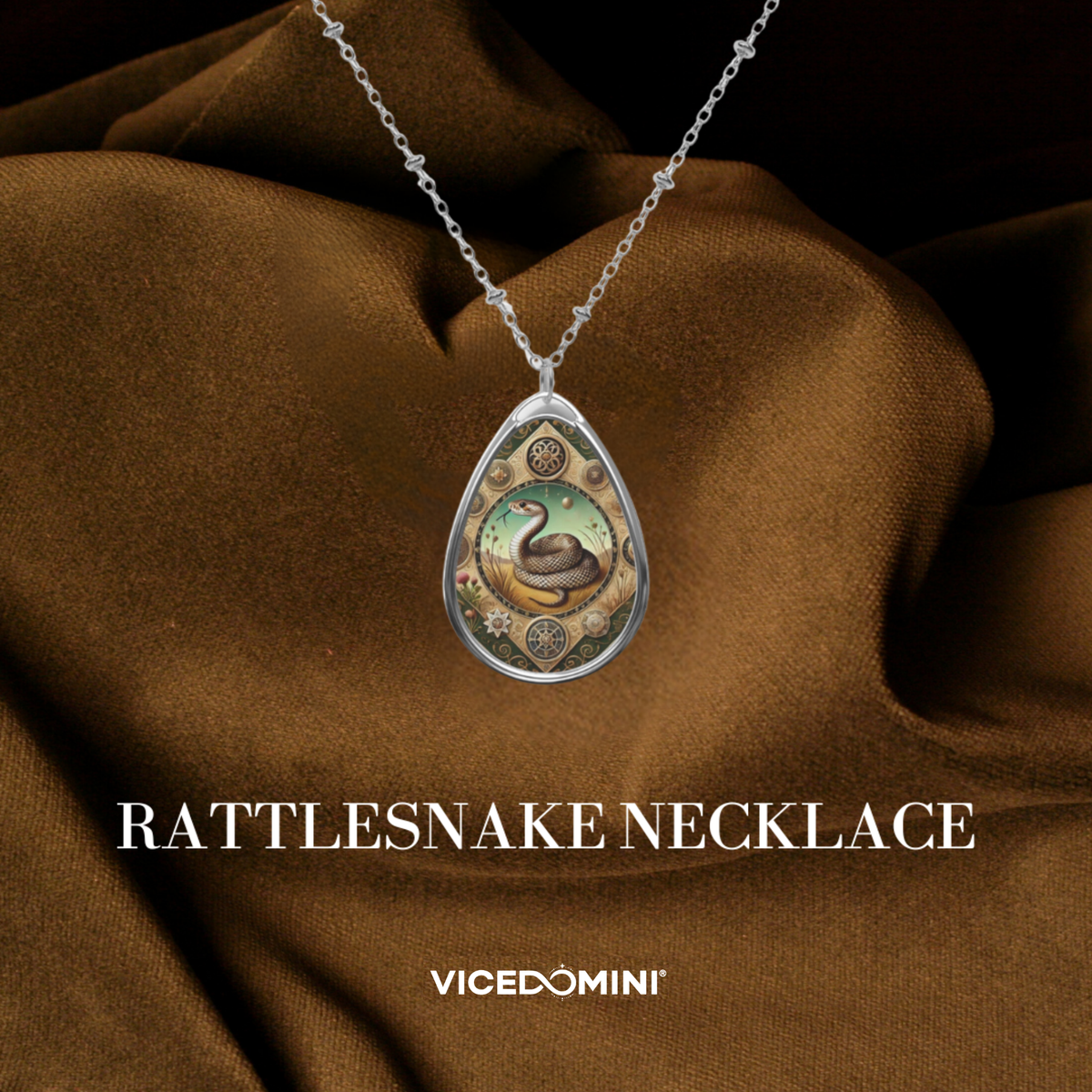 A silver teardrop pendant necklace featuring an illustrated rattlesnake design, draped over brown satin fabric with the text 'RATTLESNAKE NECKLACE' and 'VICEDOMINI' below.