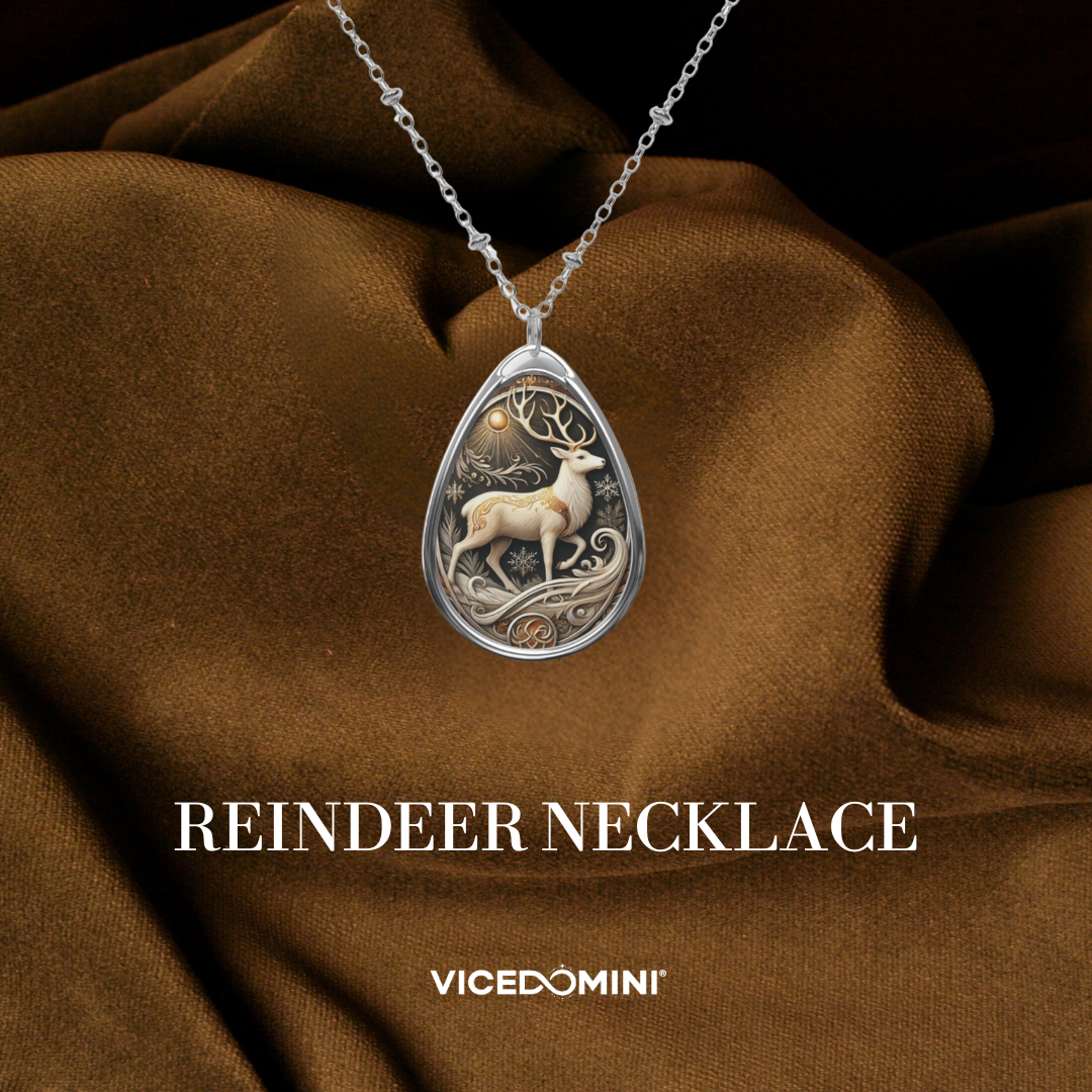 A teardrop-shaped silver pendant necklace featuring an intricate silver and gold-toned reindeer design, displayed on draped brown fabric with the text 'REINDEER NECKLACE' and 'VICEDOMINI' below.