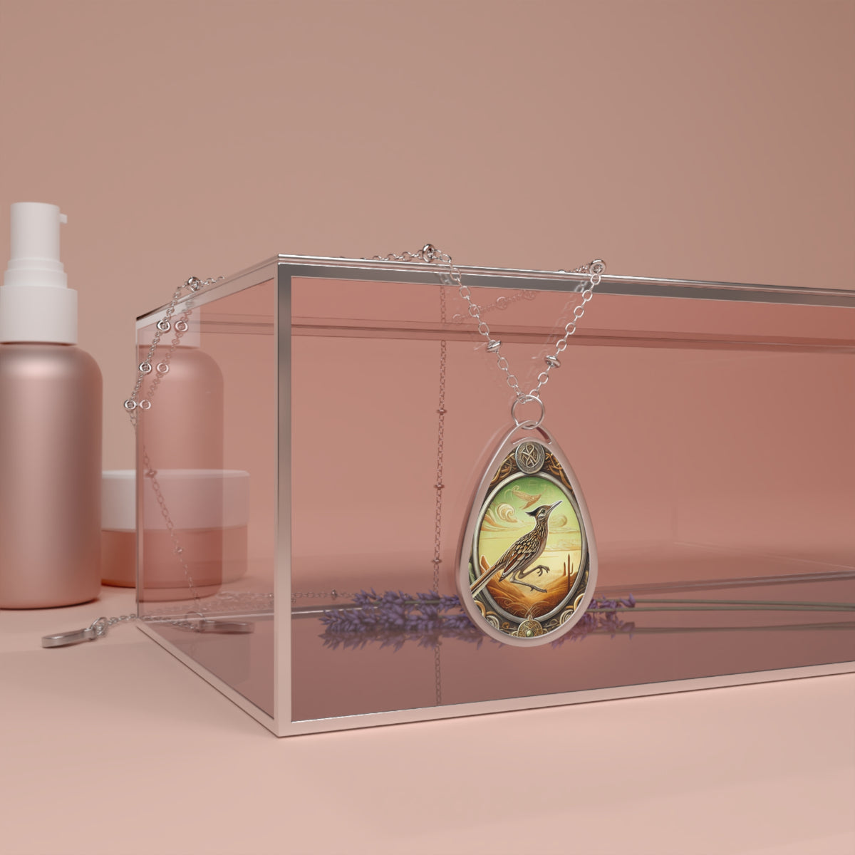 A roadrunner-themed teardrop pendant necklace displayed on a glass box with a soft pink backdrop. The necklace features intricate roadrunner artwork set against a warm desert sunset, with fine metallic detailing and a delicate chain draped elegantly.