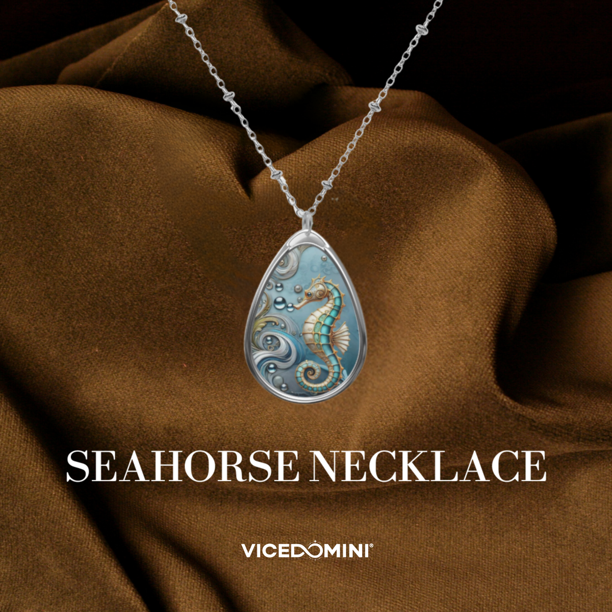 A teardrop-shaped silver pendant necklace featuring an intricate blue and gold seahorse design, displayed on draped brown fabric with the text 'SEAHORSE NECKLACE' and 'VICEDOMINI' below.