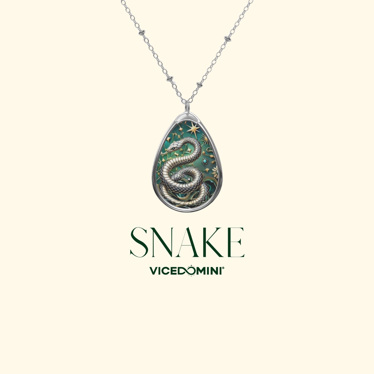 A silver-toned teardrop pendant necklace featuring an illustrated green and gold snake with stars, suspended from a delicate chain against a pale cream background. The text 'SNAKE' and 'VICEDOMINI' appears below the pendant.