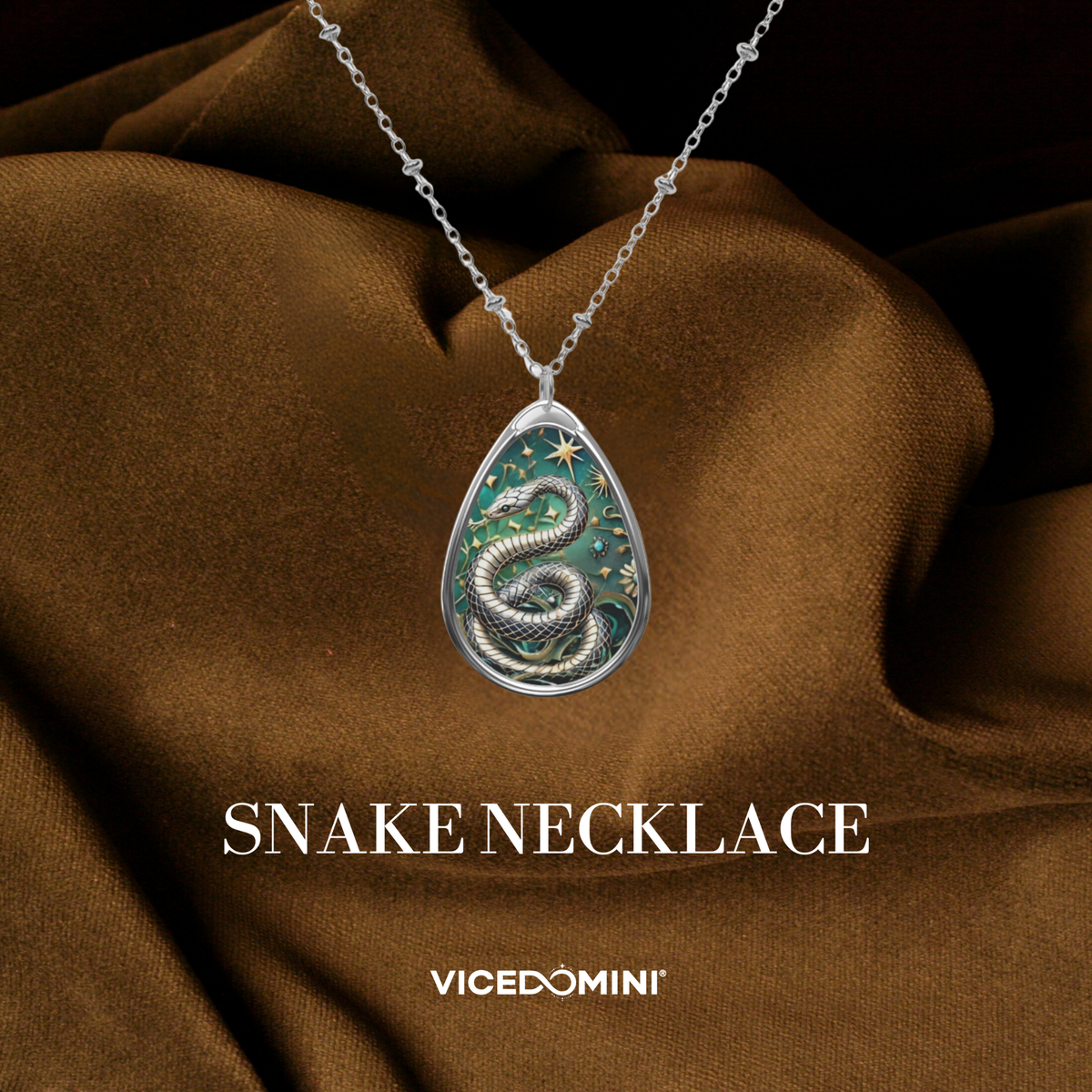 A silver teardrop pendant necklace featuring an embossed snake design over a green starry background, draped over brown satin fabric with 'SNAKE NECKLACE' and 'VICEDOMINI' text below.