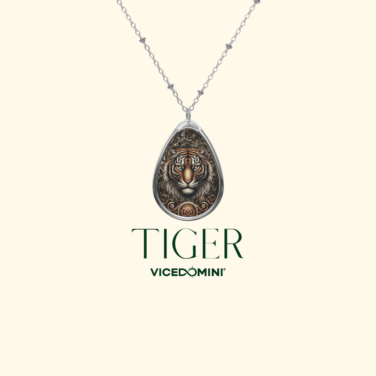 A silver teardrop pendant necklace featuring an embossed tiger face with warm metallic highlights, shown on a chain against a light cream background. Below the necklace, the brand name 'VICEDOMINI' and model name 'TIGER' are printed in dark green.