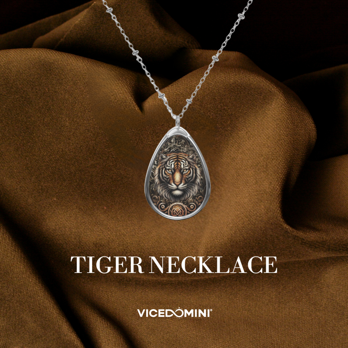 A silver teardrop pendant necklace featuring an embossed tiger face design, draped over a brown satin fabric background with the text 'TIGER NECKLACE' and 'VICEDOMINI' below.