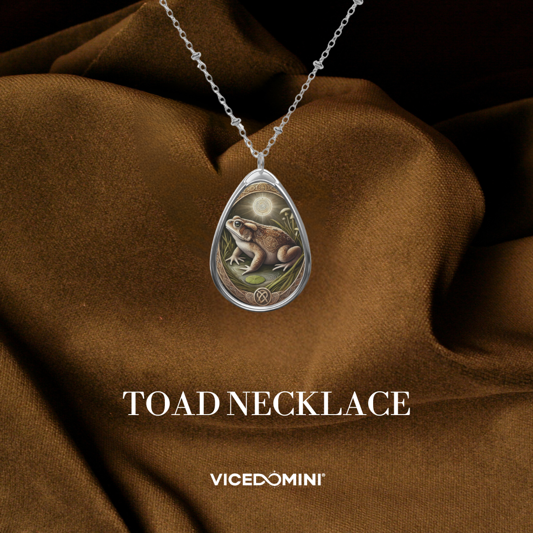 A silver teardrop pendant necklace featuring an illustration of a toad under a full moon, draped over brown satin fabric with the text 'TOAD NECKLACE' and 'VICEDOMINI' below.