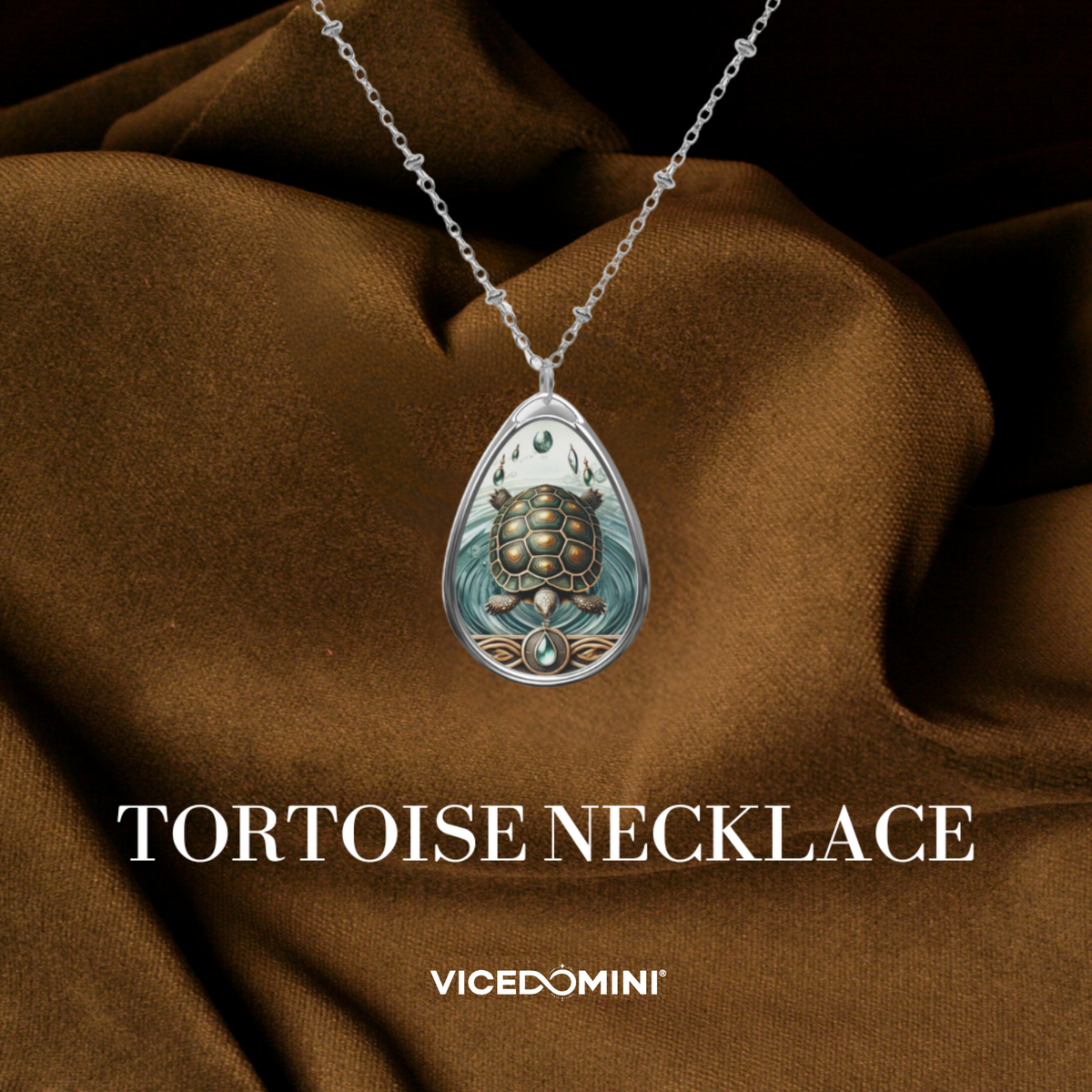 A teardrop-shaped pendant necklace with a silver-toned chain, featuring an illustrated tortoise design in muted teal, gold, and brown tones. The pendant is displayed against draped brown fabric, with the text 'TORTOISE NECKLACE' and the brand name 'VICEDOMINI' below.