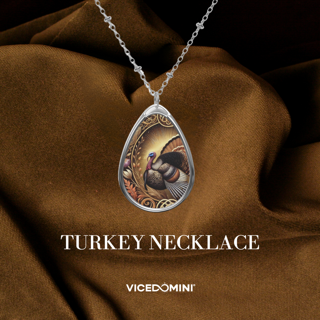 A teardrop-shaped silver pendant necklace featuring an illustrated turkey design, draped over brown satin fabric with the text 'TURKEY NECKLACE' and 'VICEDOMINI' below.