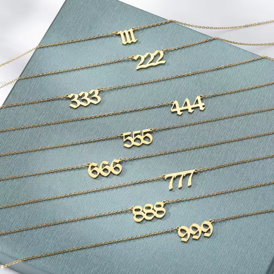 A set of gold angel number necklaces laid out on a textured blue surface, featuring numbers 111, 222, 333, 444, 555, 666, 777, 888, and 999. Each necklace has a unique font style, symbolizing differe