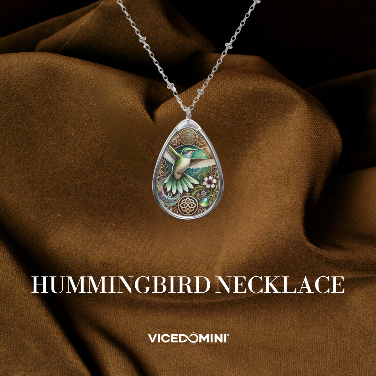 A teardrop-shaped silver pendant necklace featuring an intricate hummingbird design with green, blue, and gold details, displayed on draped brown fabric. The text 'HUMMINGBIRD NECKLACE' and the brand 'VICEDOMINI' are visible at the bottom.