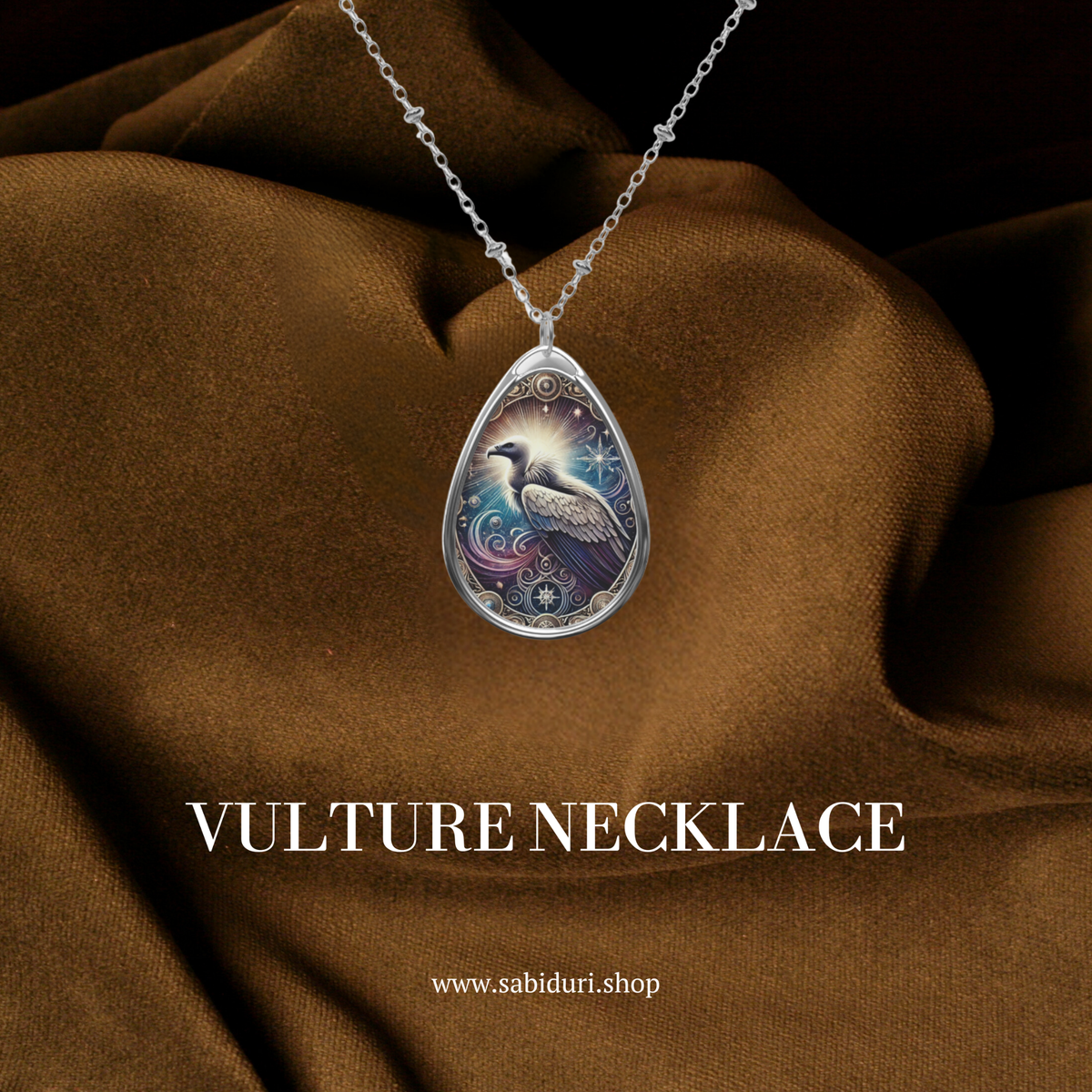 A teardrop-shaped pendant necklace featuring an intricately designed vulture illustration with radiant golden and celestial details. The pendant hangs from a delicate silver chain, displayed against a rich brown fabric background with "VULTURE NECKLACE" and "vicedomini" text overlay.