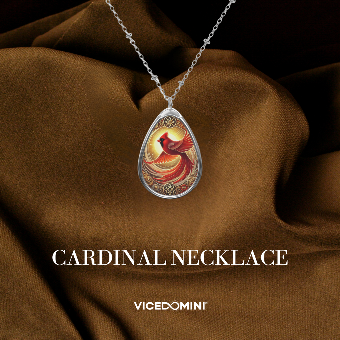 Silver teardrop pendant necklace featuring a colorful illustration of a cardinal bird in a circular frame, draped over brown satin fabric with the text 'CARDINAL NECKLACE' and 'VICEDOMINI' below.