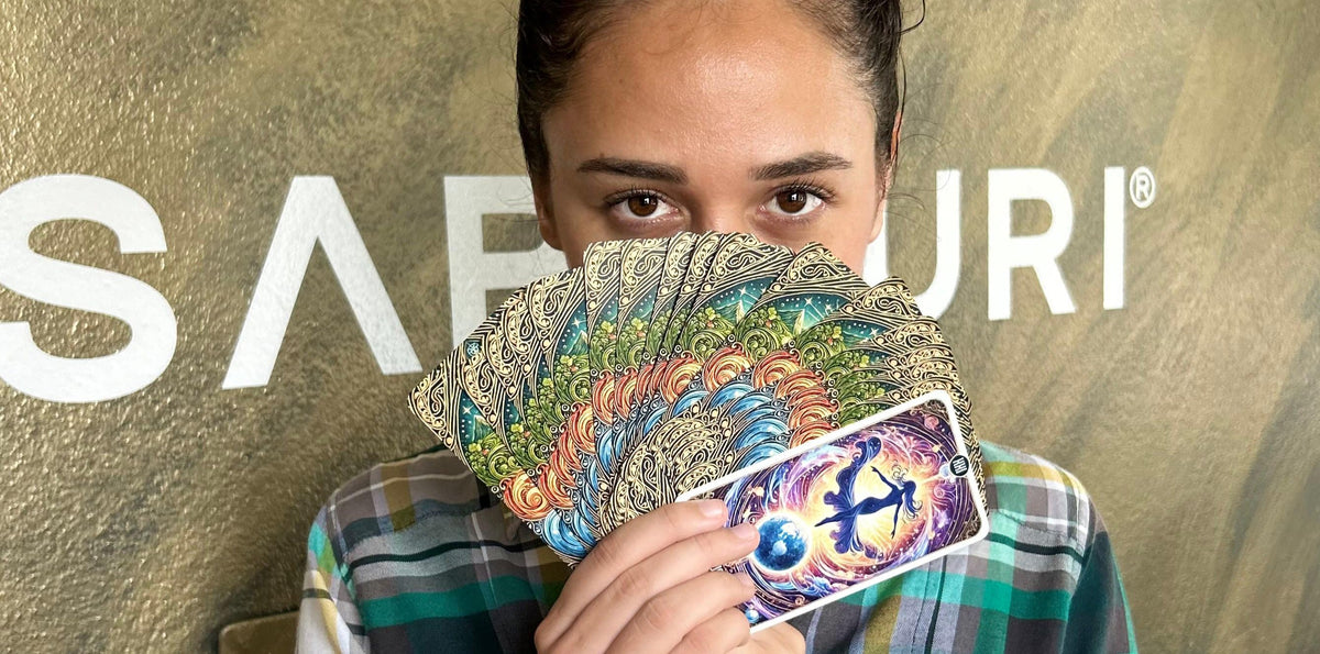 A person holds a fan of intricately designed tarot cards, mostly concealing their face except for their eyes. The tarot cards feature vibrant colors and patterns, with the card closest to the camera displaying an illustration of a cosmic figure dancing above a glowing Earth. The background shows part of a sign with the letters "SABIDURI" visible.