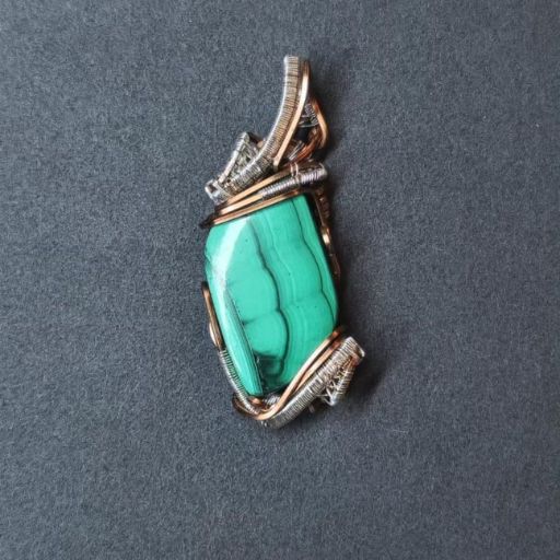 A pendant showcasing a large, bright green malachite stone with characteristic banding, wrapped in a mix of silver and copper wire, displayed on a grey surface.
